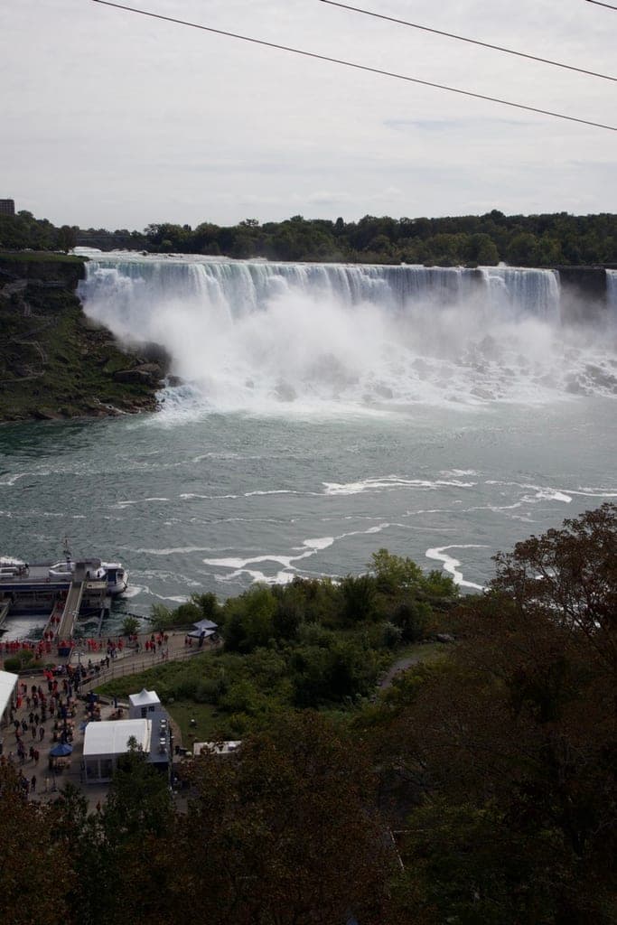 niagara's american falls from above on an overcast day, mist rising off the river, with a tour boat docked below and a crowd in red ponchos on the walkway